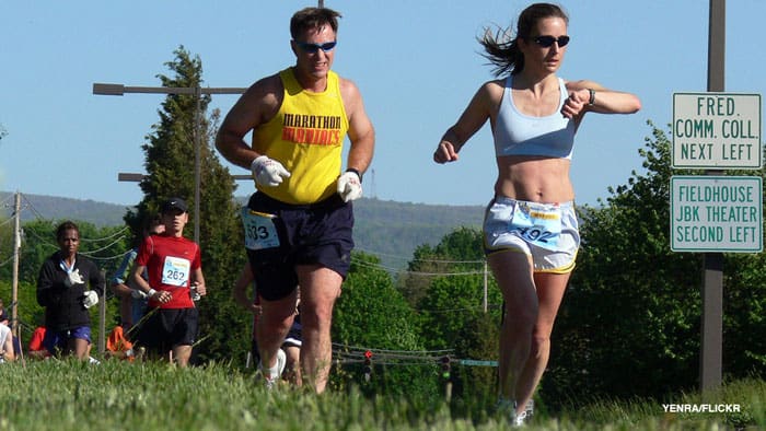 A group of people participate in an outdoor marathon on a sunny day. One woman in front checks her watch while running. A man follows closely behind, wearing a yellow tank top and gloves. Road signs can be seen in the background, guiding directions.