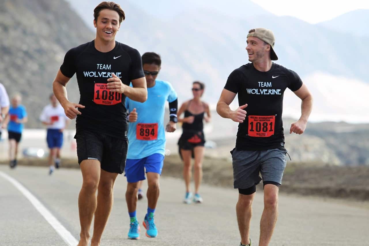 The image shows two men running a race on a road, wearing black "Team Wolverine" shirts with bib numbers 1080 and 1081. Other runners are visible behind them on a scenic route with mountains in the background. The man with bib 1081 is wearing a backward cap.