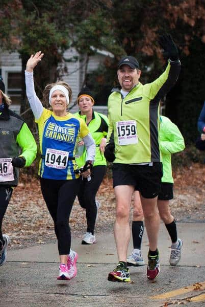 A group of runners participates in a race. Two runners in the forefront are smiling and waving at the camera. The woman on the left is wearing a yellow and blue shirt with a headband, and the man on the right is wearing a green jacket and black shorts.