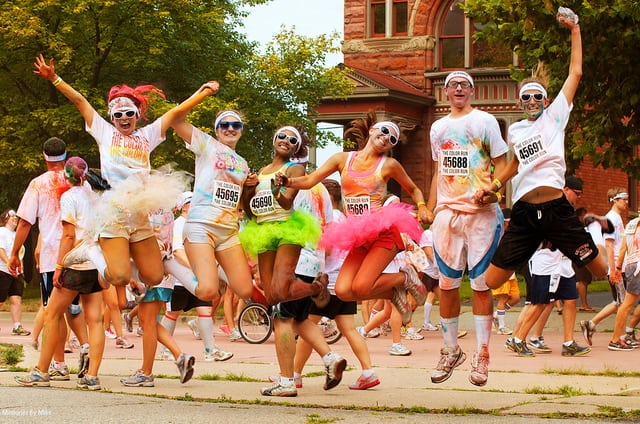 A group of runners wearing colorful tutus, sunglasses, and white shirts cheerfully jump in the air during a vibrant outdoor event. Their clothes are splashed with various colors. A large building and other participants form the lively backdrop.