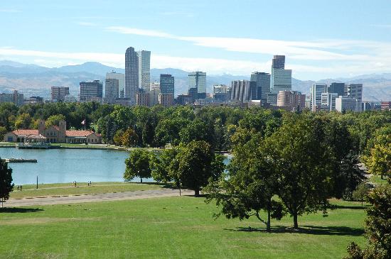 View of a city skyline with modern high-rise buildings in the background. In the foreground, there's a lush green park with trees, a calm lake, and a red-roofed building near the water's edge. The sky is clear and blue.