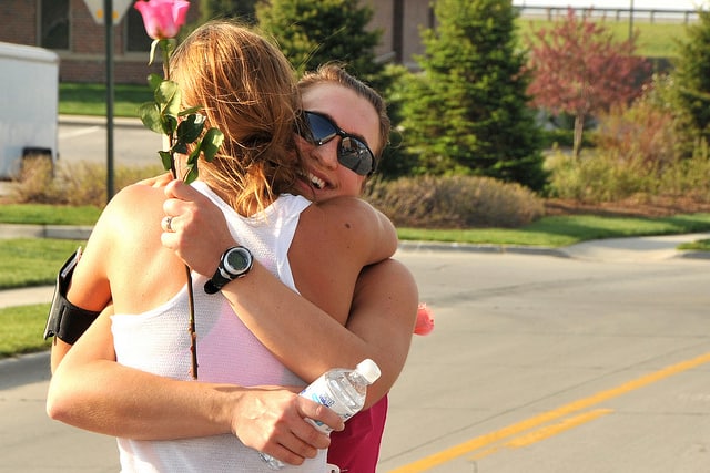 Two women embrace on a sunny day, one holding a single pink rose and the other a water bottle. They both appear happy, with one woman smiling broadly. Trees, grass, and a building are visible in the background. Both women are wearing casual, sleeveless tops.