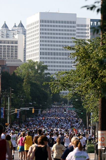 A large crowd of runners participates in a city marathon on a street lined with lush trees and tall buildings. The participants appear focused and motivated as they move towards the distance. A "NO PARKING" sign is visible on the right side of the image.