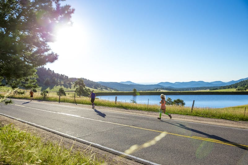 Runners on a paved road alongside a serene lake with mountains in the background. The sun is shining brightly, casting long shadows. There are green fields and a tree to the left, contributing to the scenic and peaceful atmosphere.