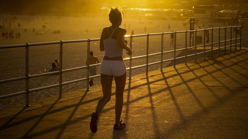 A person wearing a white tank top and shorts is jogging along a promenade during sunset. The railing casts long shadows across the ground, and beachgoers can be seen in the background enjoying the warm light and sand.