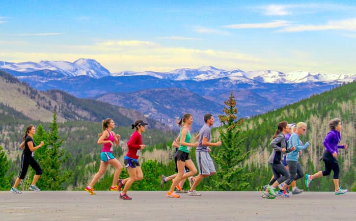 A group of people jogging on a paved road with a scenic background of lush green hills and snow-capped mountains under a blue sky with scattered clouds. The runners are dressed in colorful athletic wear.