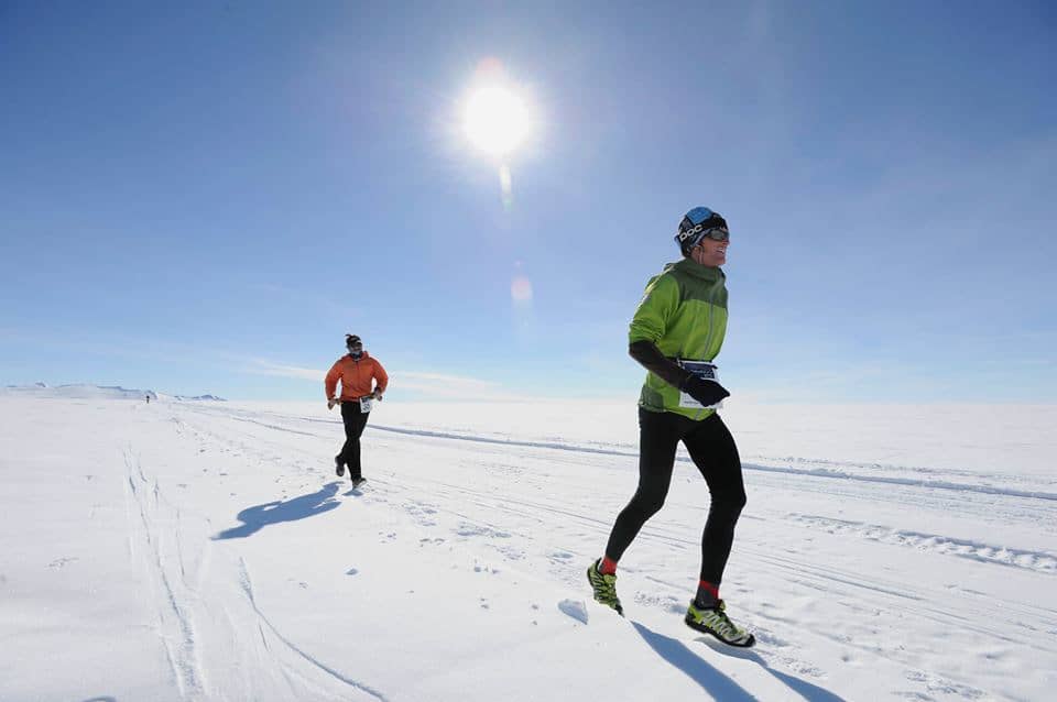 Two people running on a snowy field under a clear blue sky with the sun shining brightly. The foreground runner wears a green jacket, black leggings, and sunglasses, while the background runner wears an orange jacket and black leggings.