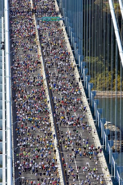 A large crowd of runners participates in a marathon, spanning both sides of a wide suspension bridge. The bridge towers and cables frame the scene, with a body of water visible on the right. The runners form a colorful, densely packed group moving forward.