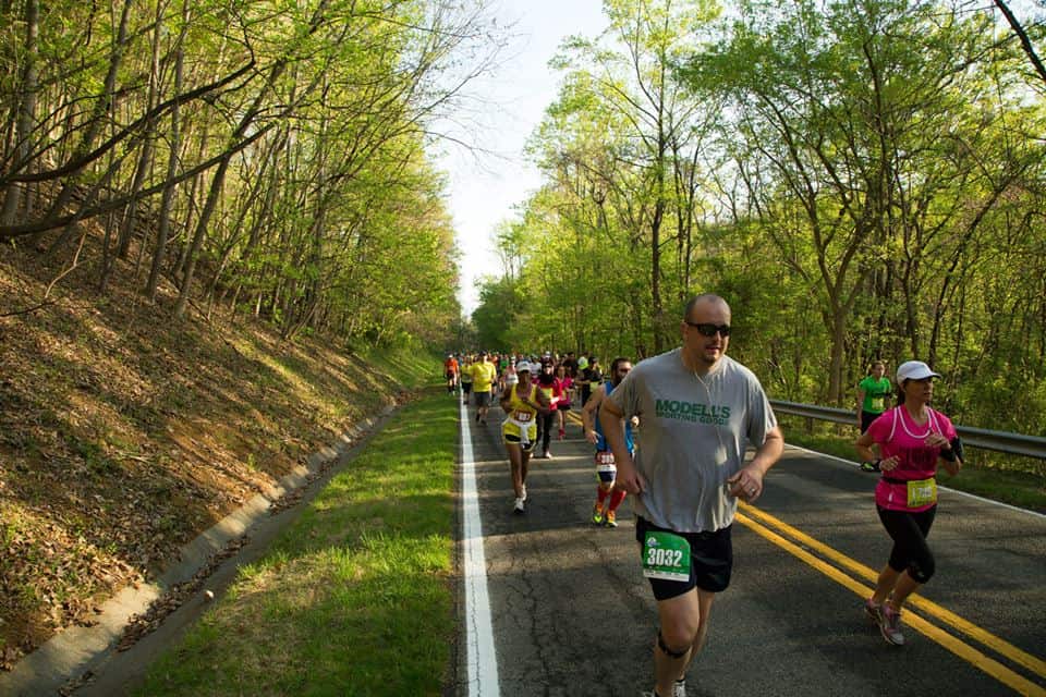 Runners participating in a race on a two-lane road surrounded by trees with fresh green leaves. Participants wear different colored clothing and race bibs. A man in a gray shirt and sunglasses is in the foreground, while others follow behind him on the course.