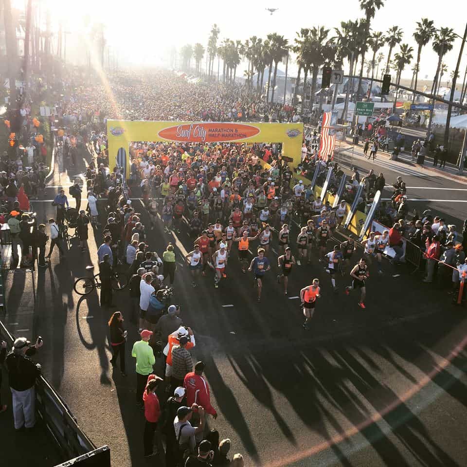 An overhead view of a large group of runners at the starting line of the Surf City Marathon. The scene is bustling with participants and onlookers. The marathon takes place near the beach, with palm trees lining the area on a bright sunny day.