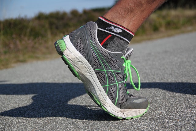 Close-up of a person's leg wearing a gray running shoe with green accents and black socks. The person is running on a paved road, and the background is slightly blurred, showing some outdoor greenery.