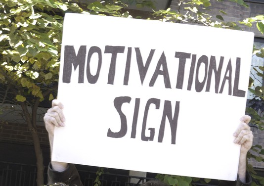 A person holds a large white sign with "MOTIVATIONAL SIGN" written in bold, black letters. The background features green foliage and part of a building.