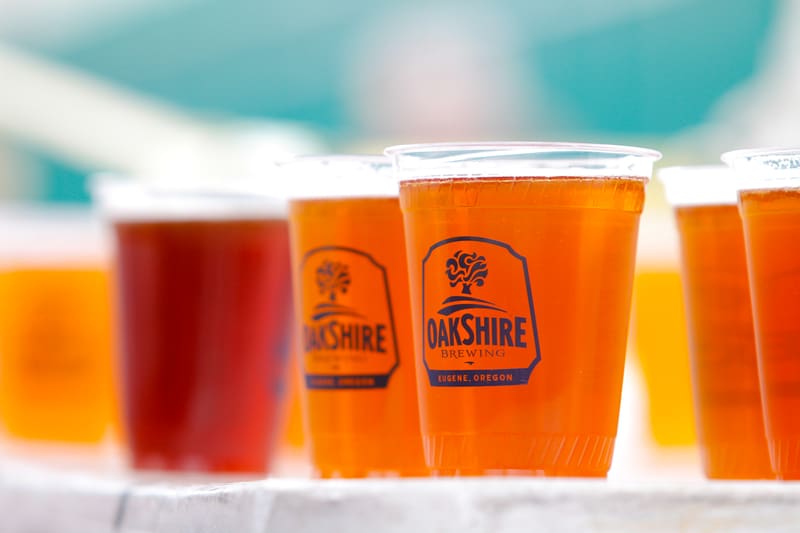 A row of clear plastic cups filled with different shades of amber-colored beer from Oakshire Brewing. The cups are placed on a white table. The Oakshire Brewing logo and text "Eugene, Oregon" are visible on the cups.