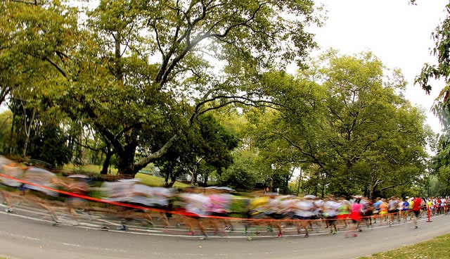 A blurred image of numerous runners participating in a marathon or race. The scene is set in a tree-lined park with tall, green trees in the background. Red tape or a line is visible, marking the path the runners are following. The runners are in motion, creating a sense of speed and dynamism in the photograph.