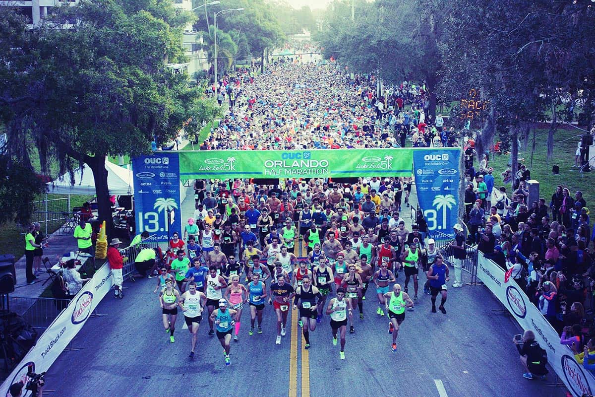 A large crowd of runners starts a race at the OUC Orlando Half Marathon. The runners are spread across a wide street, some wearing colorful attire. Spectators line the sides, and green trees are visible in the background. A banner overhead marks the start line.