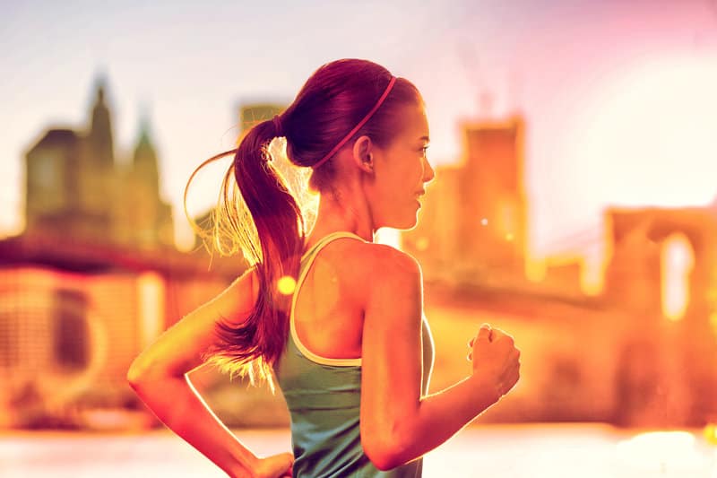 A woman with a ponytail is running outside. She is wearing a sleeveless top. The city skyline is blurred in the background with tall buildings, and the sun is shining brightly, casting a warm glow over the scene.