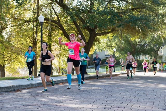 Runners participate in an outdoor race on a sunny day. A woman in a bright pink shirt and blue knee-high socks is at the forefront, smiling and gesturing energetically. Others runners follow behind her on the tree-lined, brick-paved road.