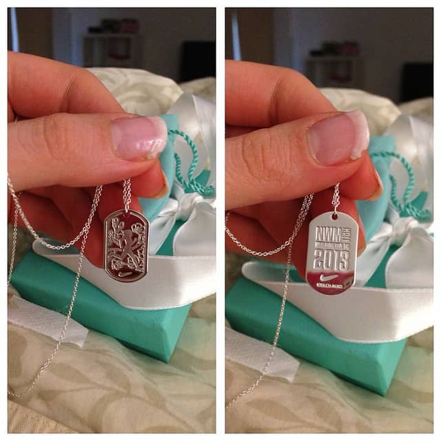 A person holds up a silver dog tag necklace. The front of the tag has an intricate design, and the back is engraved with the details: "NWM 2013" and "Nike Women's Half Marathon San Francisco." The box it came in is teal with a white ribbon.