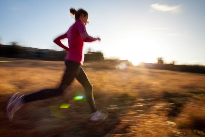 A woman in a red long-sleeve shirt and black pants is running through a field at sunrise or sunset. The image is slightly blurred to show motion, and there is a warm, glowing light in the background.