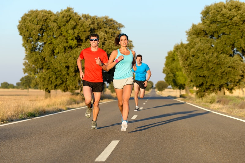 Three people are running on a paved road surrounded by nature. The runners, wearing athletic clothes, are focused and appear to be enjoying their exercise. Tall trees and dry grass line the sides of the road, and the sky is clear with sunlight.