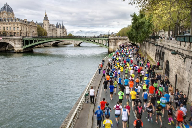 Hundreds of runners participate in a marathon along the Seine River in Paris. They run on a riverside path with a historic bridge in the background and buildings and trees lining the route. The scene is vibrant, with runners wearing colorful outfits.