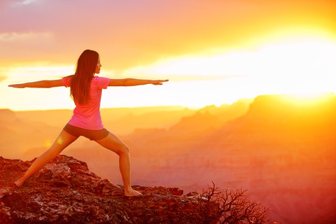 A woman performs a yoga pose outdoors on a rocky cliff at sunrise or sunset. She is standing in a Warrior II stance, with arms extended, facing the sun that creates a warm, golden glow over the landscape. The background shows a vast, mountainous terrain.