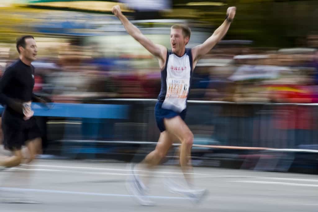 A male runner in athletic attire and a race bib crosses the finish line with arms raised in victory, while another runner in black follows behind. The background is blurred, emphasizing their movement and the speed of the race.