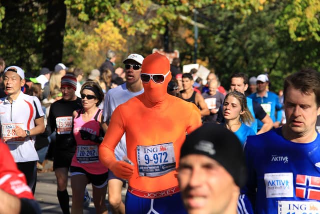 A group of marathon runners in various athletic gear runs along a sunlit path lined with trees and spectators. One runner stands out in a full-body orange suit and white sunglasses, drawing attention among the diverse crowd. Race bibs display “ING” and individual numbers.