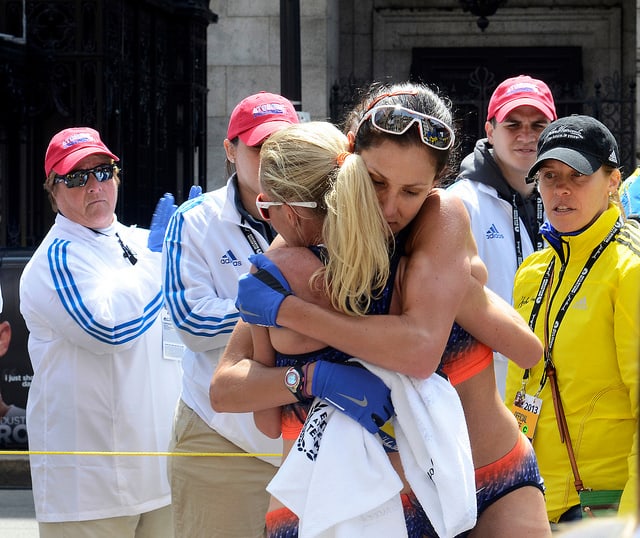 Two athletes embrace warmly after a race, wearing sports gear and blue gloves. Their emotional moment is witnessed by four officials in white uniforms with red hats and one in a yellow jacket. A building with a dark entrance is visible in the background.