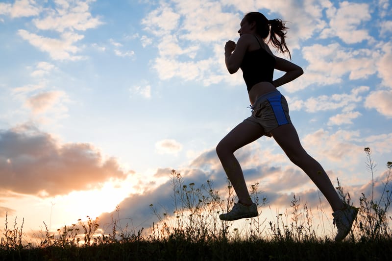 A person is shown running outdoors in silhouette against a picturesque sky with colorful clouds at either sunrise or sunset. They are dressed in athletic wear, including a tank top, shorts, and running shoes. The sunlight creates a dramatic and serene atmosphere.