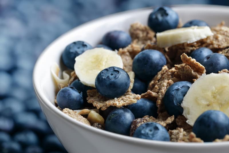 A close-up of a white bowl filled with cereal flakes, fresh blueberries, and banana slices. The cereal appears textured, and the fruits add vibrant color. The background is blurred but shows more blueberries scattered around.