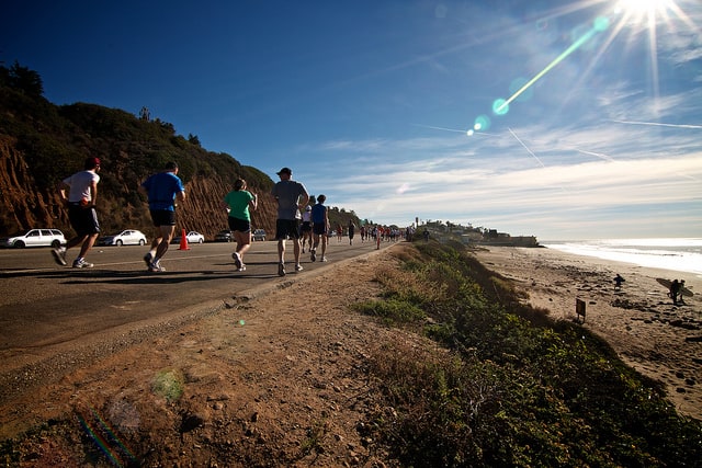 A group of people jogging on a paved road beside a cliff and the ocean under a bright, sunny sky with a clear view of the coastline. Some cars are parked on the side of the road, and the sun is creating a lens flare in the image.