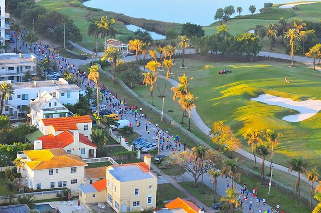 Aerial view of a marathon as runners move along a road lined with palm trees. On one side of the road, colorful houses and buildings are seen, while on the other side is a green golf course with a few trees and sand traps. The background reveals a body of water.