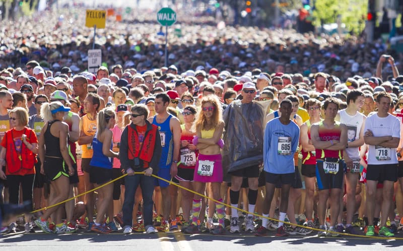 A large crowd of runners, dressed in colorful athletic gear, stands at the starting line of a marathon. Some runners wear race bibs and there is a yellow rope marking the start. Signs in the background indicate walking and race path directions.