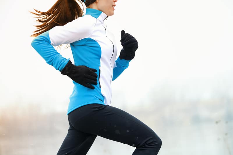 A person dressed in a blue and white jacket, black pants, and black gloves is running outdoors. The background is an overcast sky, suggesting a cool or cold day. The runner's ponytail is seen swinging with their motion.