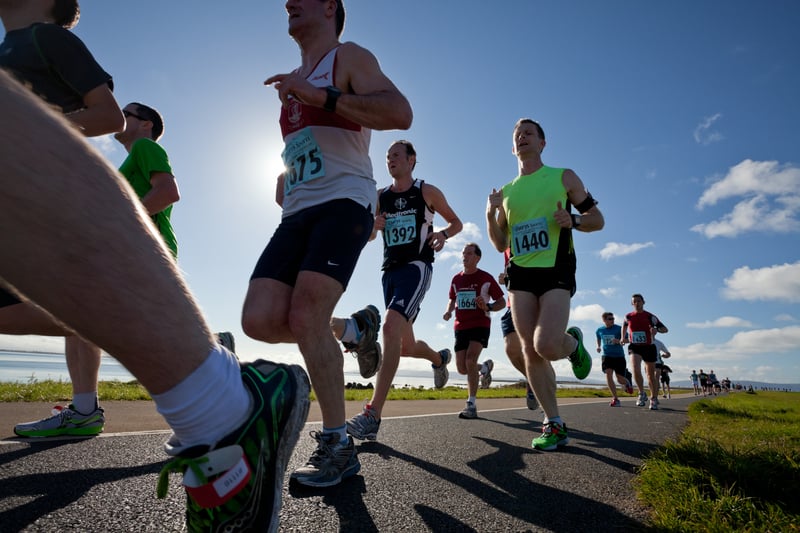 A group of runners participating in a race on a sunny day, with some wearing race numbers and athletic gear. They are running on a paved road near a grassy area with blue skies and scattered clouds overhead. A coastal view is visible in the background.