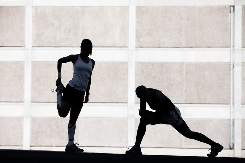 Silhouettes of two athletes stretching against a grid-like wall. The person on the left is holding one leg behind them, while the person on the right is in a lunging position. Both are dressed in athletic wear and appear to be warming up.