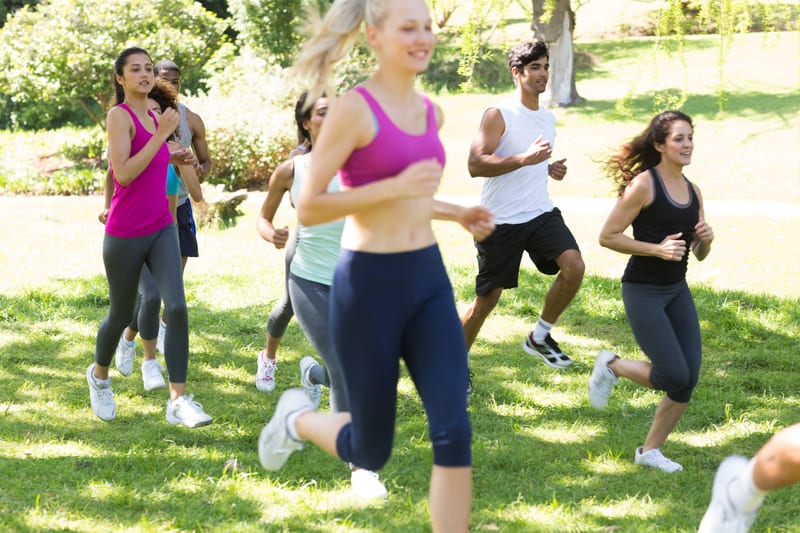 A group of people jogs in a park on a sunny day. They are wearing athletic clothing, and there are trees and greenery in the background. The individuals appear to be of various ages and are engaged in a fitness activity.