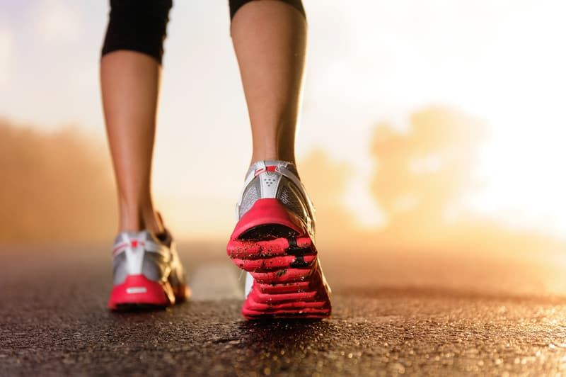 Close-up of a person walking or running on a paved surface at sunrise or sunset. The focus is on their lower legs and red and white sneakers, which are in motion. The background is blurred, creating a vibrant and dynamic feel.