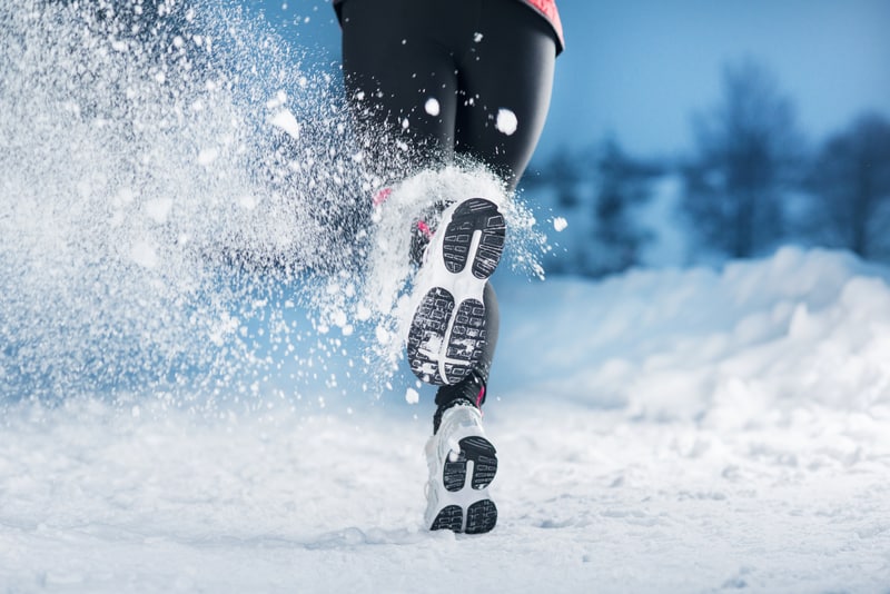 A person in winter running gear runs through snow, causing snow to spray up from their feet. The ground and surroundings are covered in snow, and the sky appears to be overcast. This dynamic shot captures the movement and energy of a winter run.