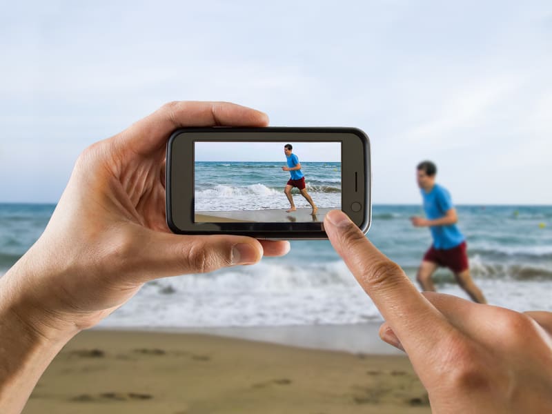 A person is photographing another person running on the beach via a smartphone. In the smartphone's screen, the runner is visible in the frame with the ocean in the background. The beach scene includes sand, waves, and a clear sky.