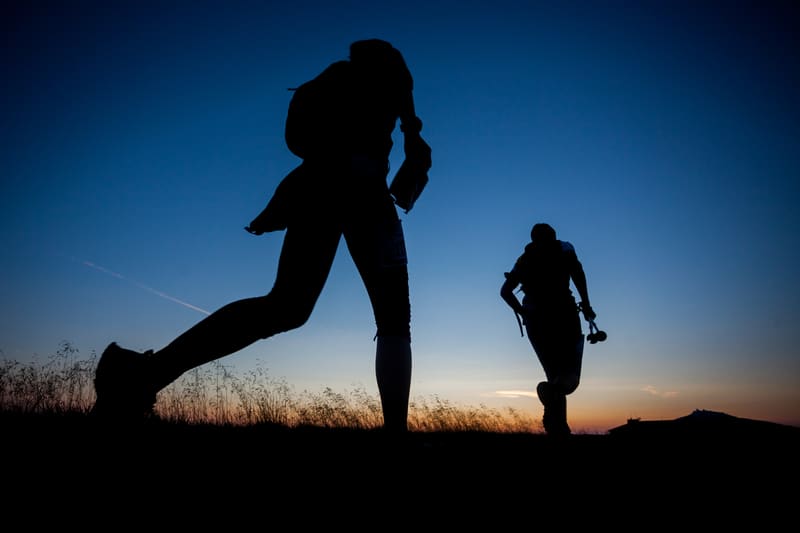 Silhouettes of two hikers walking on a grassy hill at dusk. The sky is a gradient of deep blue fading into orange near the horizon. One hiker in the foreground appears to be in mid-stride, while the other is slightly behind and to the right.