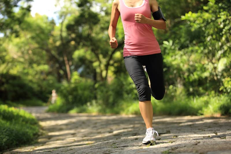 A woman jogs on a stone path through a lush, green park. She is wearing a pink tank top, black leggings, and white running shoes. An armband is visible on her left arm. The background features tall trees and foliage.