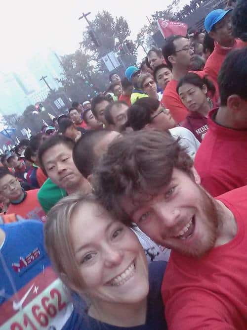 A large group of participants in a marathon race are gathered together, smiling and excited. The front of the crowd features a man with a beard and a woman with blonde hair, both grinning at the camera. Other runners are seen in the background, wearing colorful shirts.