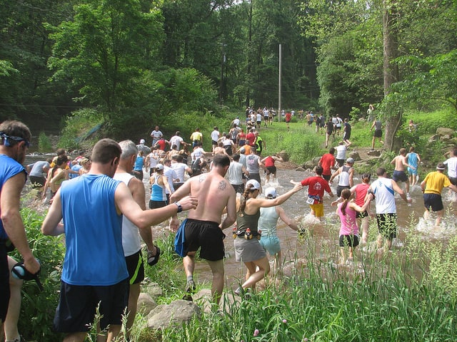 A large group of people, wearing athletic clothing, are participating in a race through a forested area. They are wading through a shallow creek, with lush greenery and trees surrounding the path. The runners are helping each other across the water.