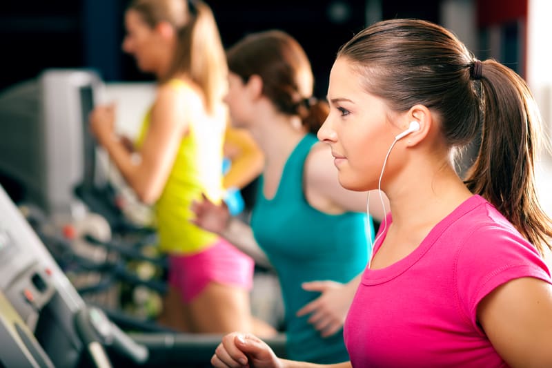 Women are running on treadmills at a gym. In the foreground, one woman wears a pink shirt and has earbuds in, focusing ahead. Other women in bright workout clothes are visible in the background, exercising on adjacent treadmills.