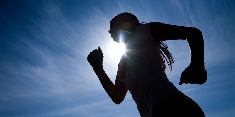 Silhouette of a person running against a bright sun and a blue sky. The sunlight creates a halo effect around their head and shoulder, emphasizing the motion and athleticism. The figure's hair is tied back, suggesting they are likely engaged in a vigorous run or jog.