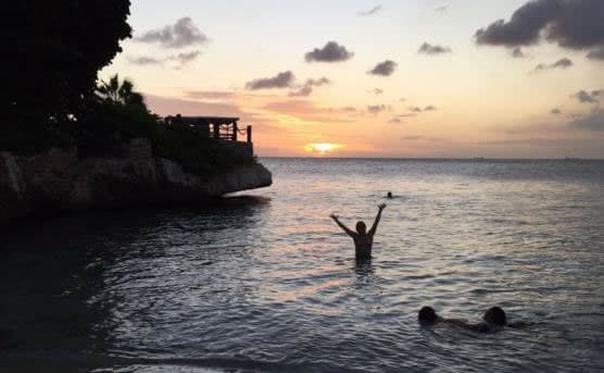 A person stands waist-deep in the sea with their arms raised, enjoying the sunset. Two other people are swimming nearby. The sun dips below the horizon, casting an orange glow over the water, while trees and a structure silhouette against the sky on the left.