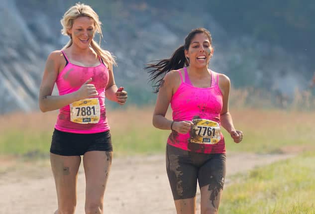 Two women, wearing pink tops, participate in an outdoor race. Both have mud on their clothes and bodies, indicating a challenging course. They wear race bibs with numbers 7881 and 7761. The background shows a dirt path with greenery on the sides.