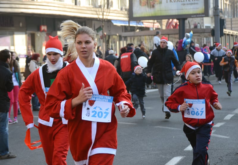 People dressed in Santa Claus outfits are running in a street race. The participants, including women, men, and children, wear red suits and hats. Some spectators are watching from the sidewalk. It appears to be a festive event during the holiday season.